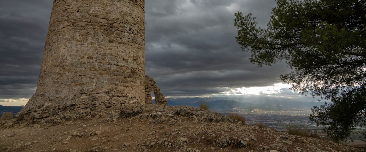 A stone tower with a large hole in the middle, surrounded by rocky terrain and a cloudy sky.