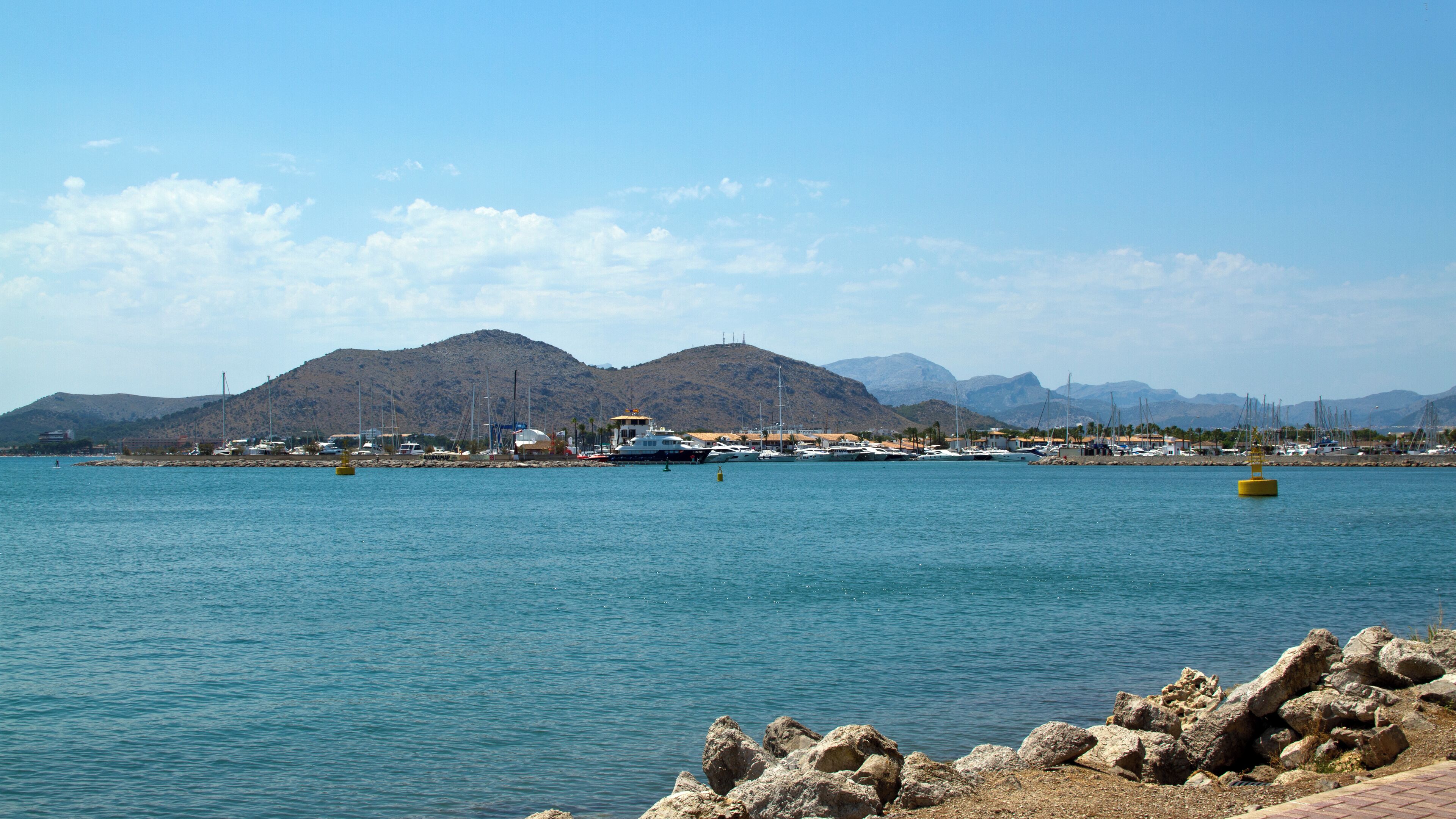 A coastal marina with boats, rocky shoreline, and mountains in the background.
