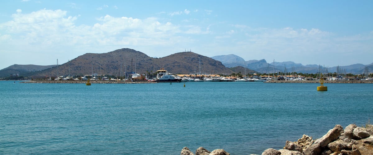 A coastal marina with boats, rocky shoreline, and mountains in the background.
