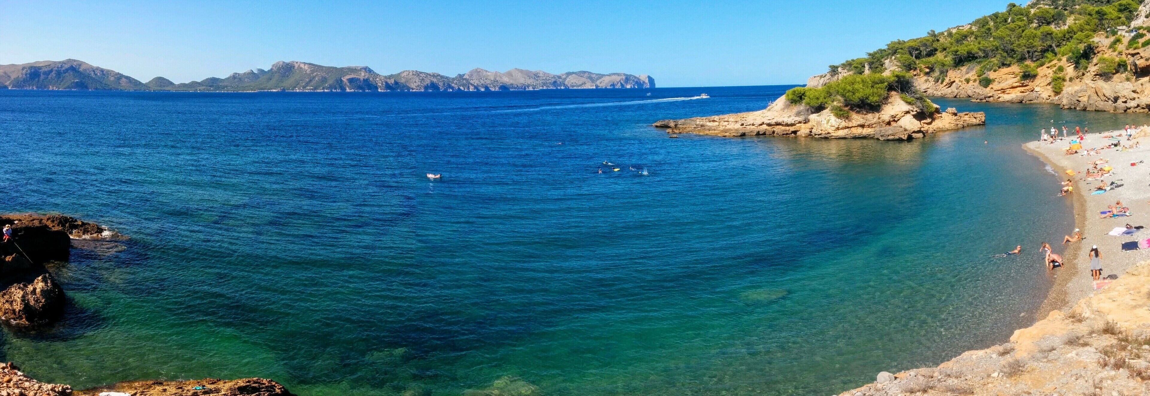 A coastal beach with clear blue waters, rocky outcrops, and distant mountains.