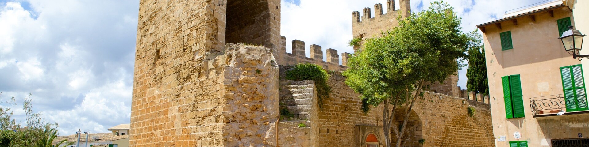 A historic stone tower with arched openings and battlements, surrounded by buildings with green shutters.