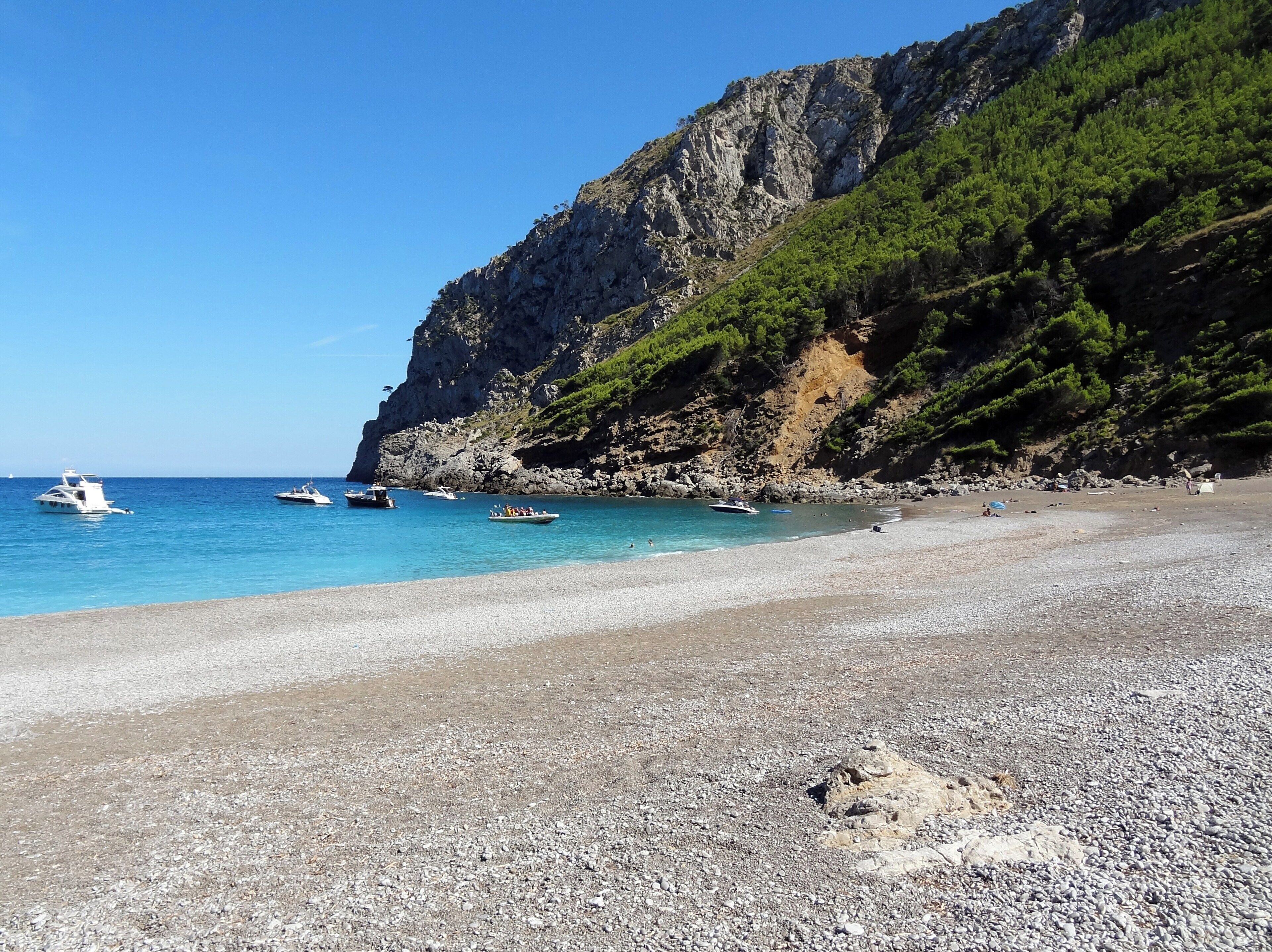 A pebble beach with boats, a rocky cliff, and clear blue water.