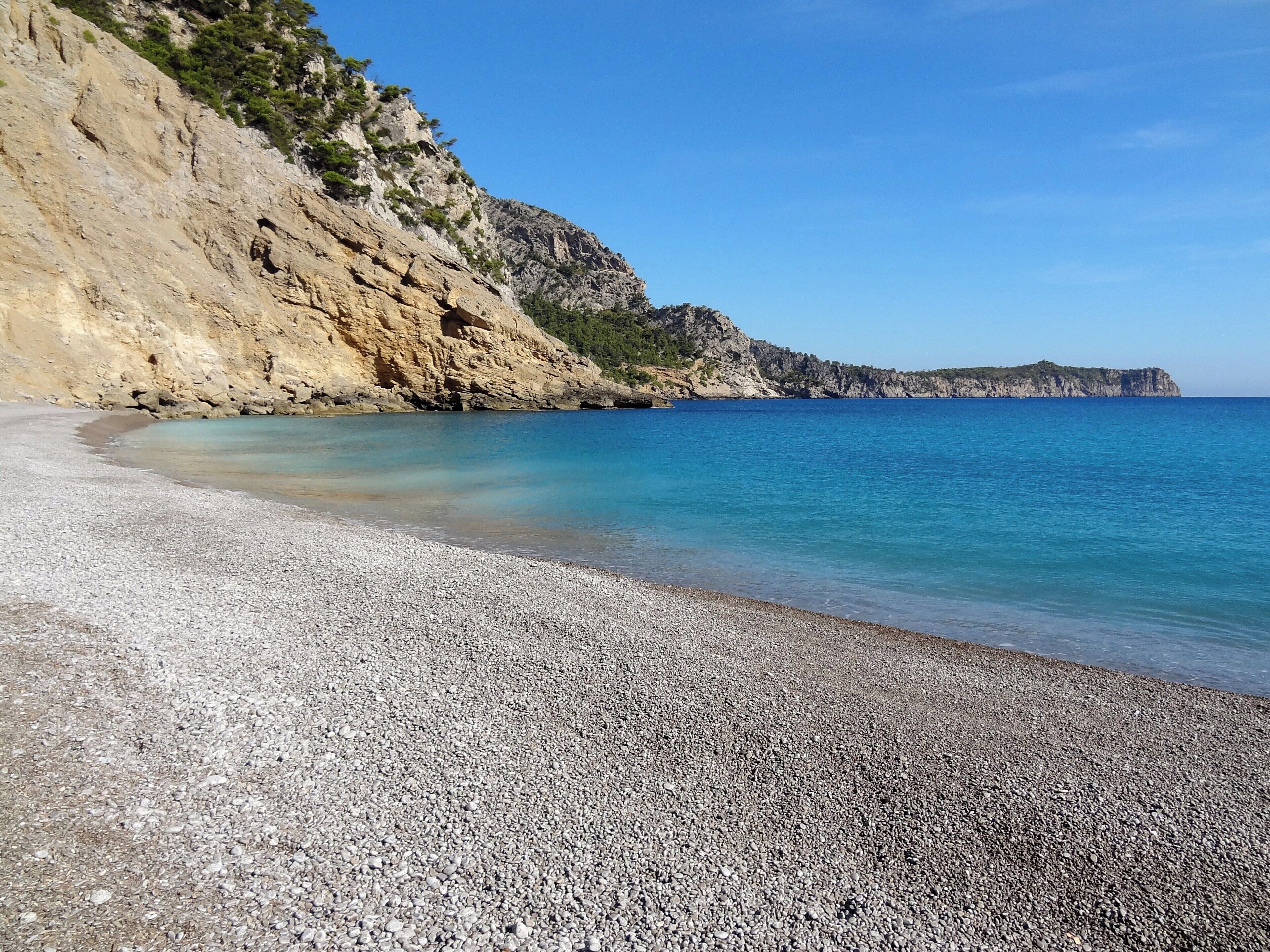 A pebble beach with clear turquoise water and rocky cliffs.