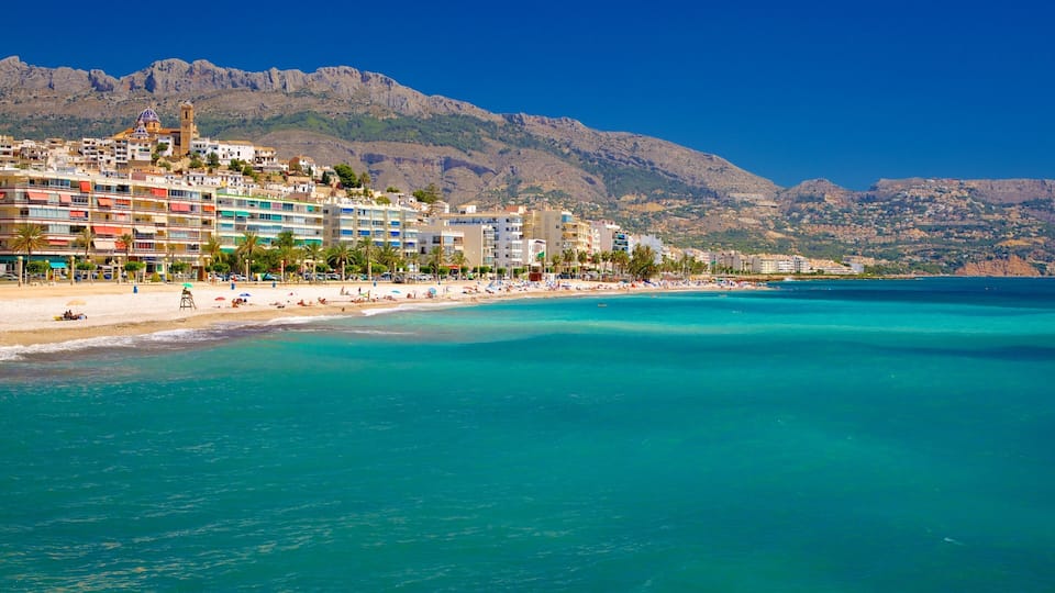 A coastal town with a beach, clear turquoise waters, and mountains in the background.