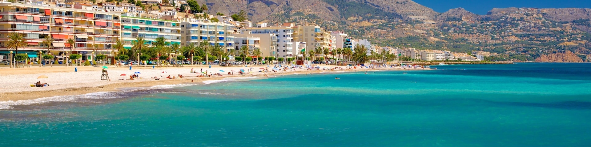 Un pueblo costero con una playa, aguas turquesas cristalinas y montañas al fondo.