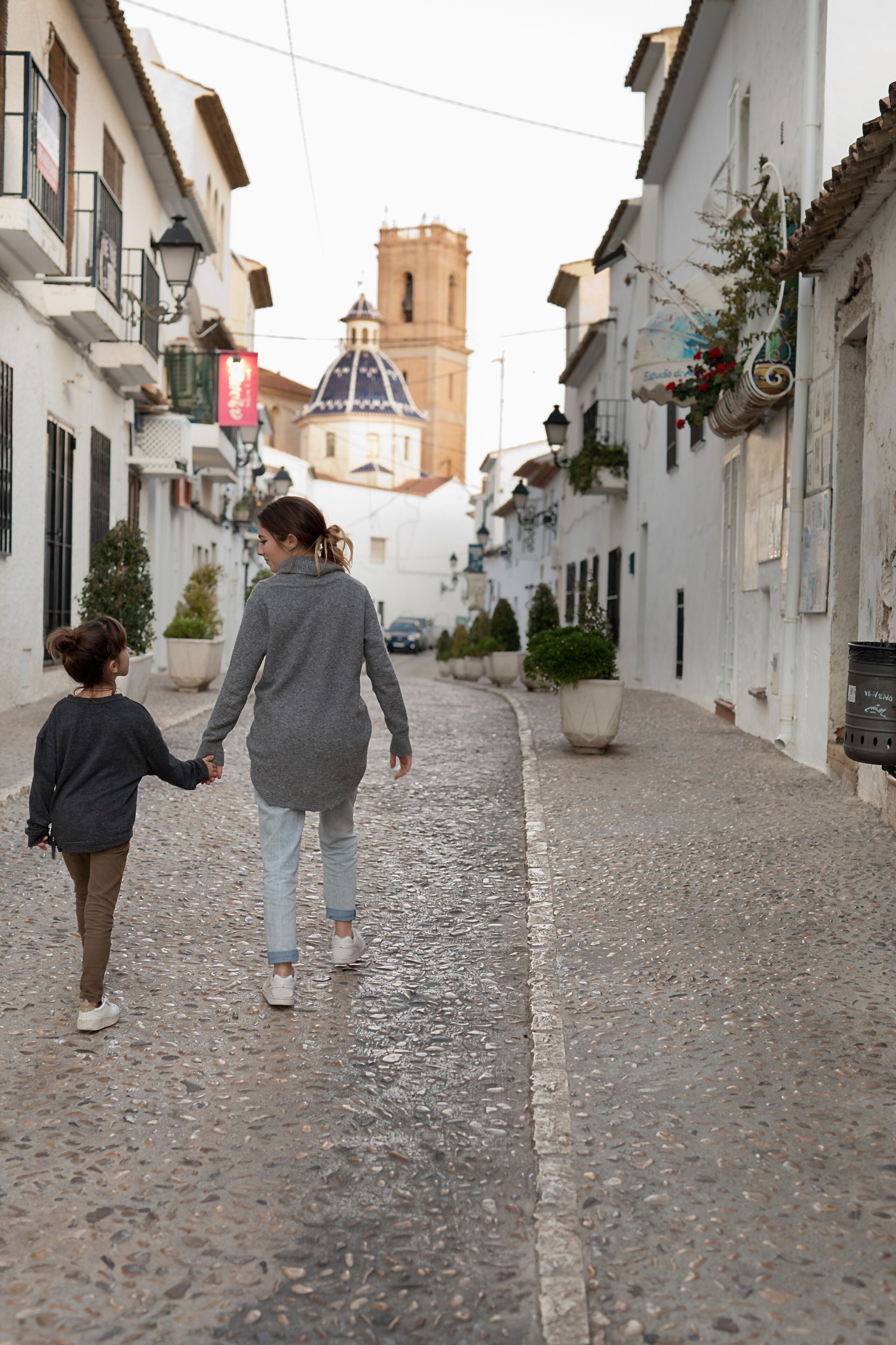 Una calle adoquinada con edificios, una torre de iglesia y macetas colgantes con flores.