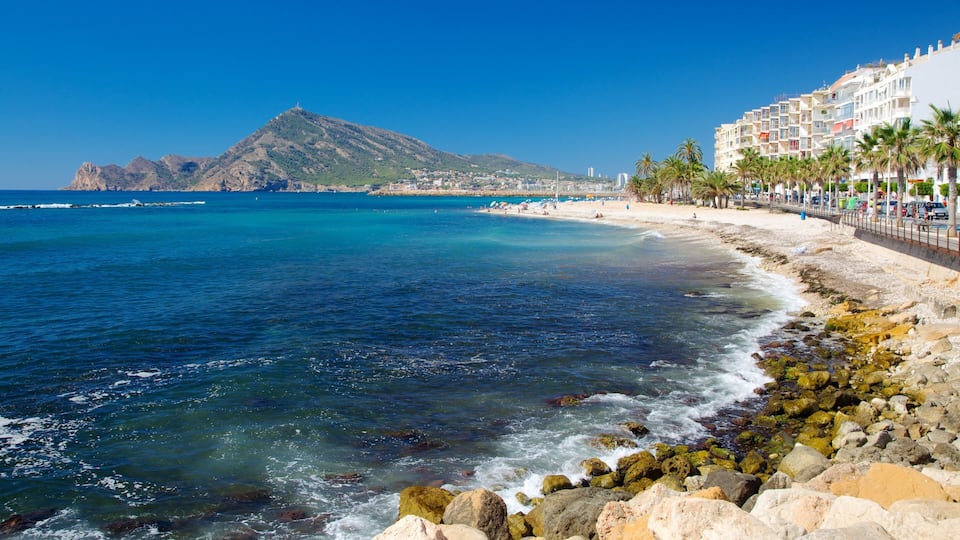 A coastal beach with clear blue waters, rocky shoreline, and a mountain in the background.