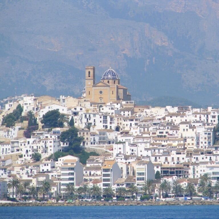 A coastal town with a prominent church featuring a dome and bell tower, surrounded by densely packed buildings and palm trees.