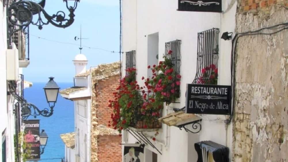 A narrow street with white buildings, a restaurant sign, and hanging flower baskets.