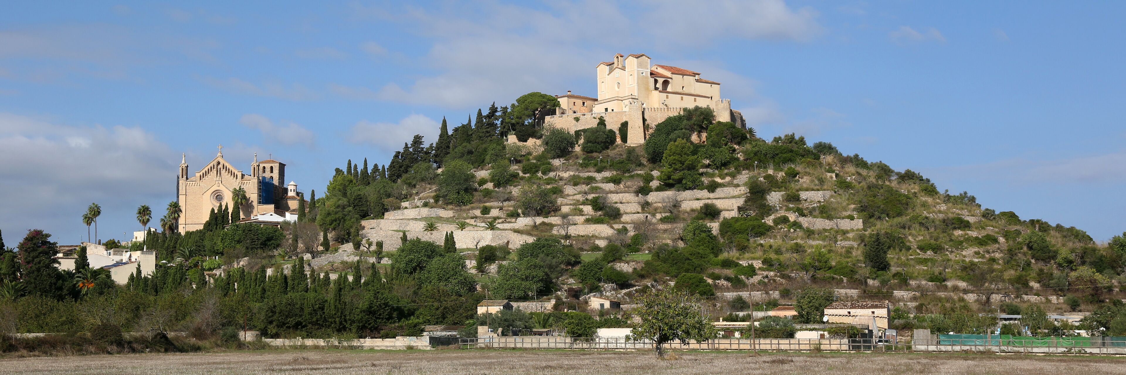 A hilltop church with a bell tower and a castle-like structure on a hill with trees and a clear sky.