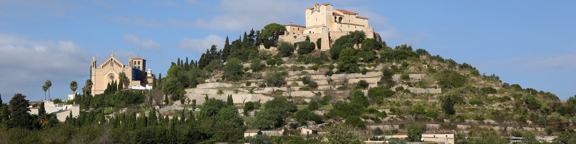 A hilltop church with a bell tower and a castle-like structure on a hill with trees and a clear sky.