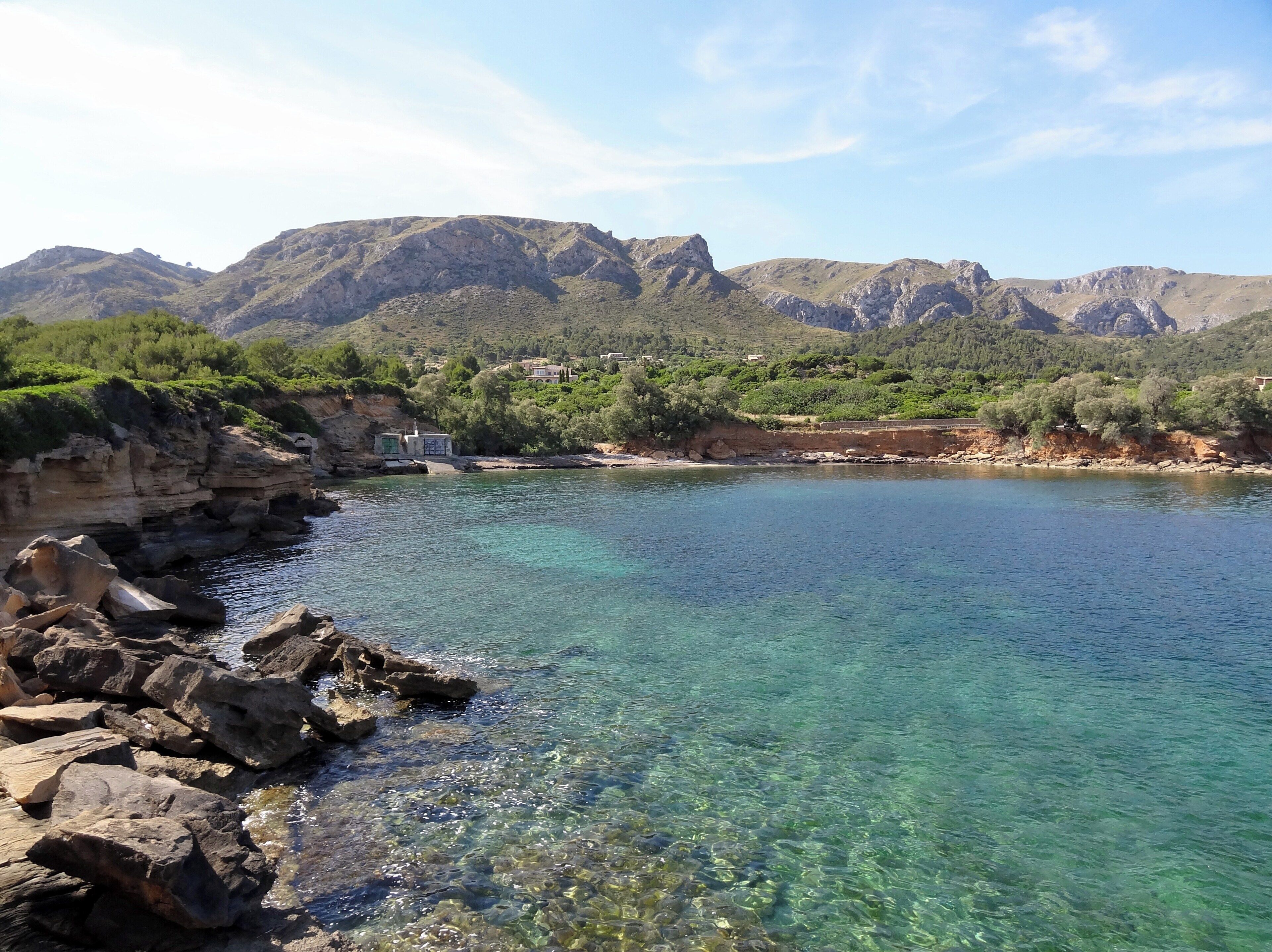 A coastal landscape with clear turquoise waters, rocky shores, and mountainous terrain in the background.