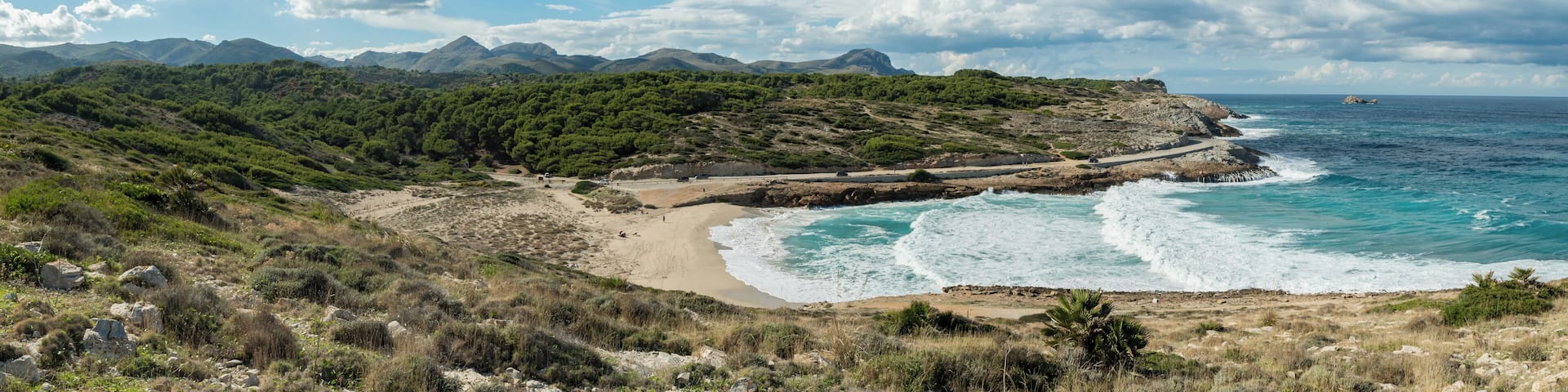 A coastal landscape with a sandy beach, rocky cliffs, and a mountainous backdrop.