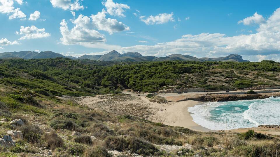 A coastal landscape with a sandy beach, rocky cliffs, and a mountainous backdrop.