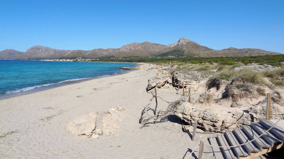A beach with clear blue water, sandy shore, and mountains in the background.