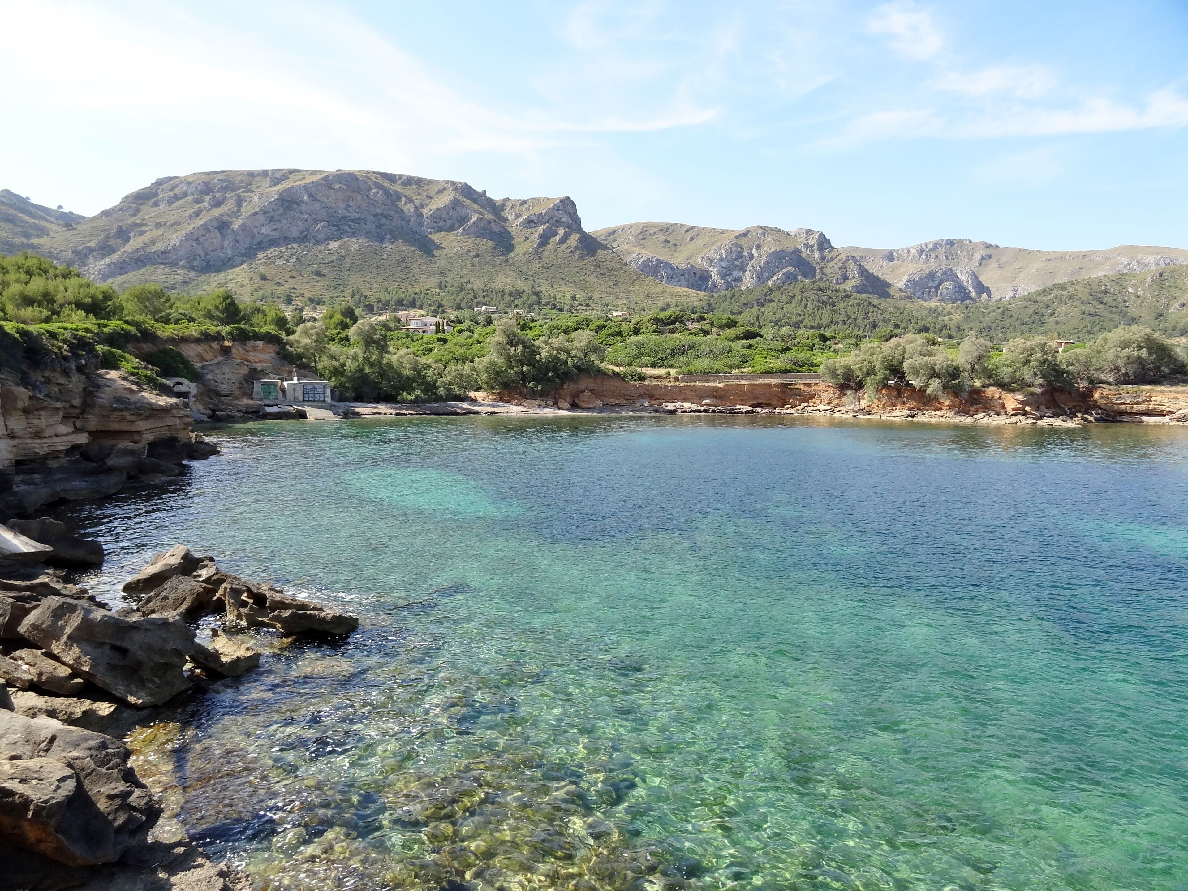 A clear coastal area with rocky shores, a calm sea, and mountains in the background.