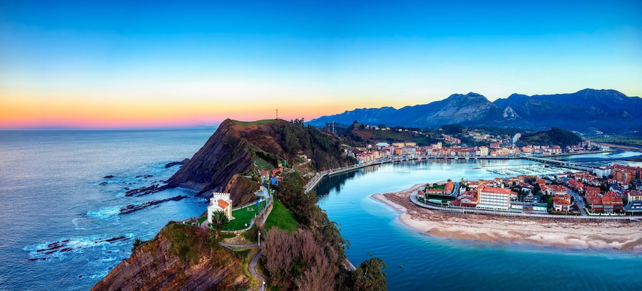 Uma cidade costeira com um penhasco imponente, um porto de recreio e uma cordilheira ao fundo.