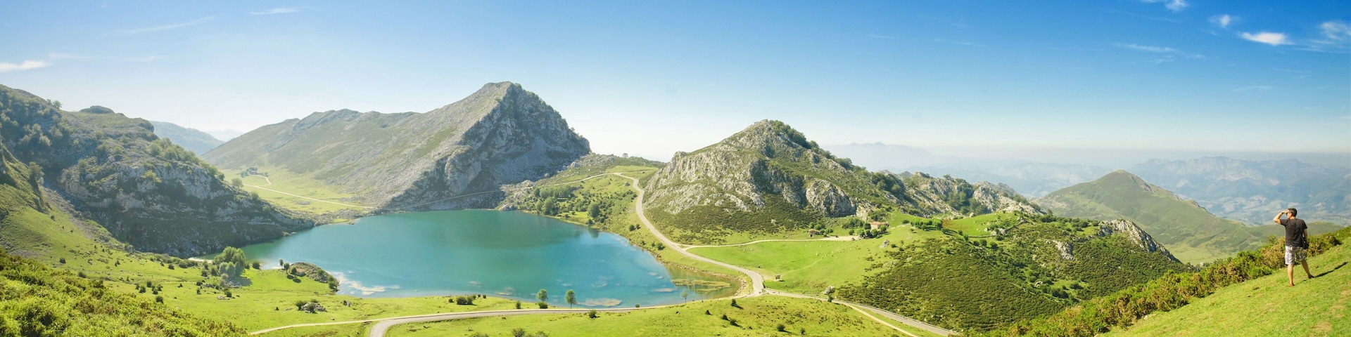 Paisaje montañoso con un lago, carreteras sinuosas y una persona de pie en una colina.