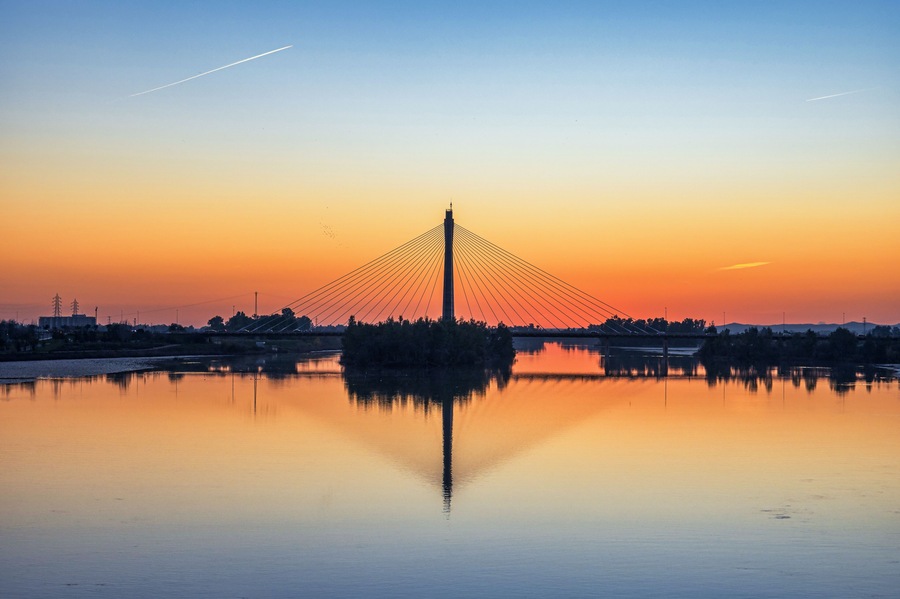 En kabelbro i solnedgang med en rolig flod der spejler broens konstruktion.