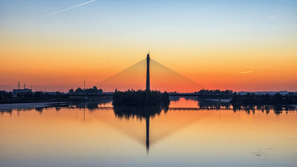 A cable-stayed bridge at sunset with a calm river reflecting its structure.