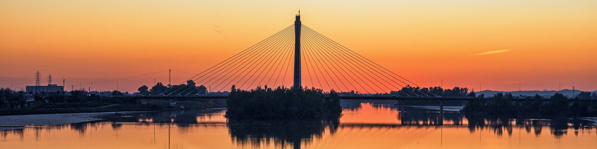 En kabelbro i solnedgang med en rolig flod der spejler broens konstruktion.
