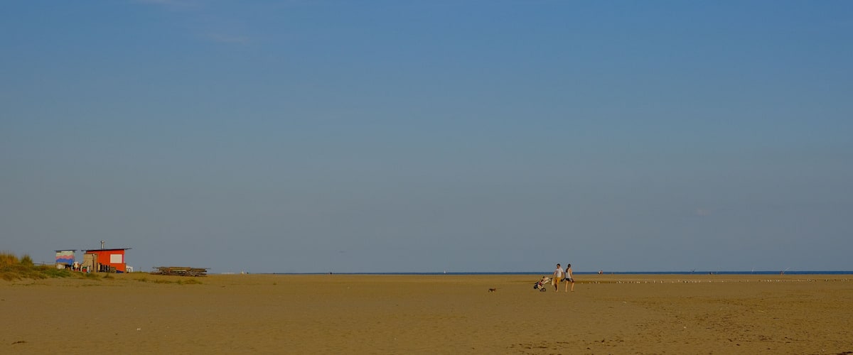 A wide sandy beach with a few structures, including a red building and a small shed, under a clear blue sky.