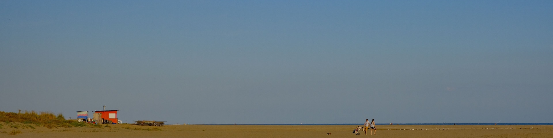 A wide sandy beach with a few structures, including a red building and a small shed, under a clear blue sky.