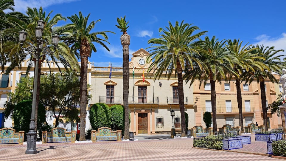 A plaza with palm trees, a clock tower, and a building with balconies.