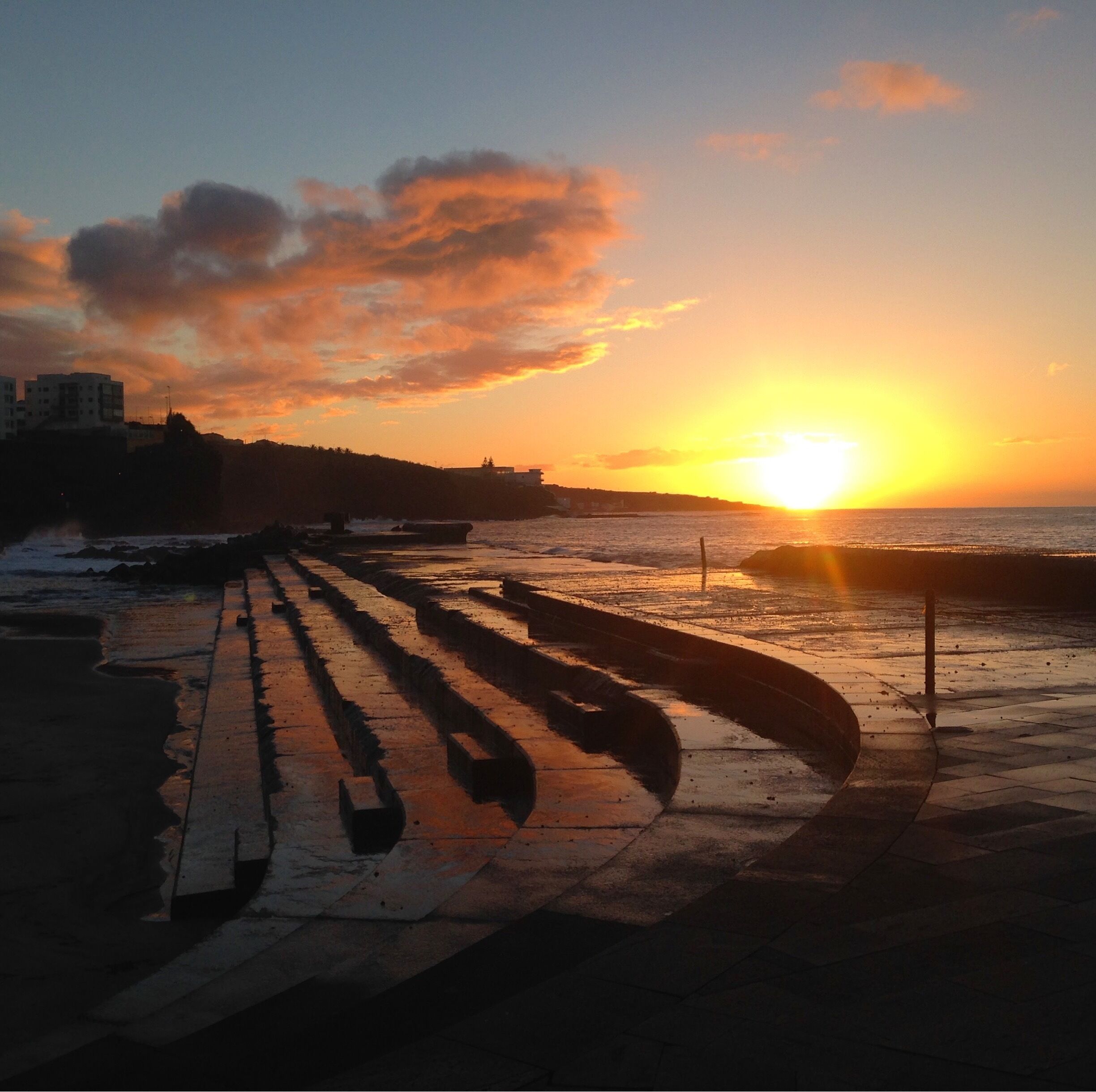 A sunset over a coastal promenade with a boardwalk and buildings in the background.