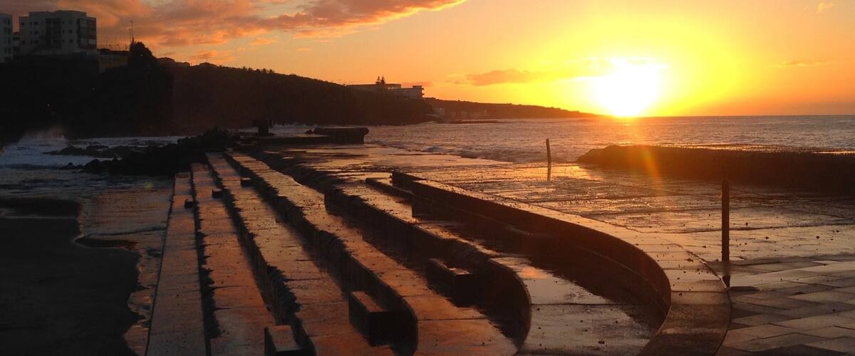 A sunset over a coastal promenade with a boardwalk and buildings in the background.