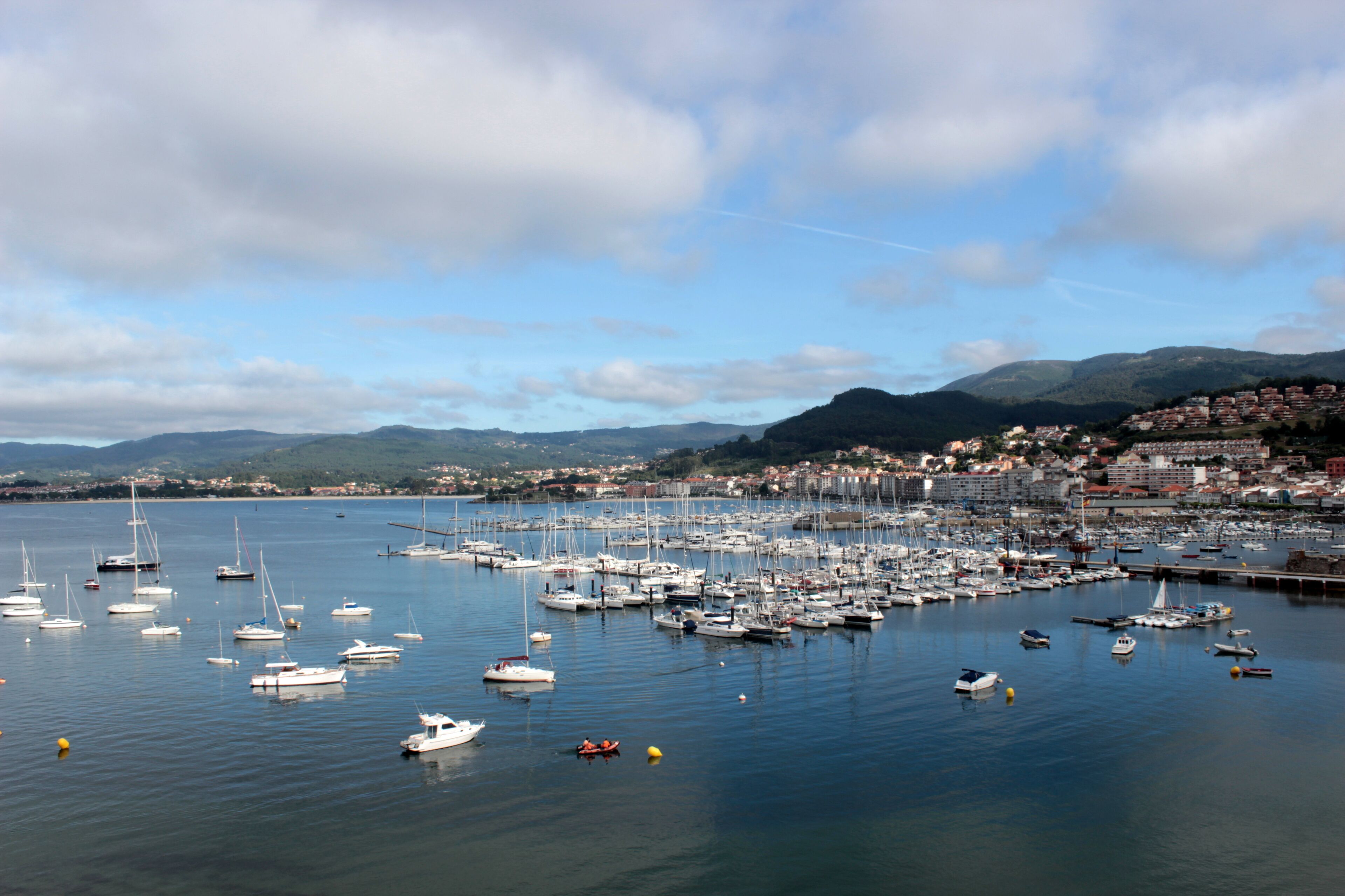 A marina with numerous boats docked, a coastal town in the background, and mountains in the distance.