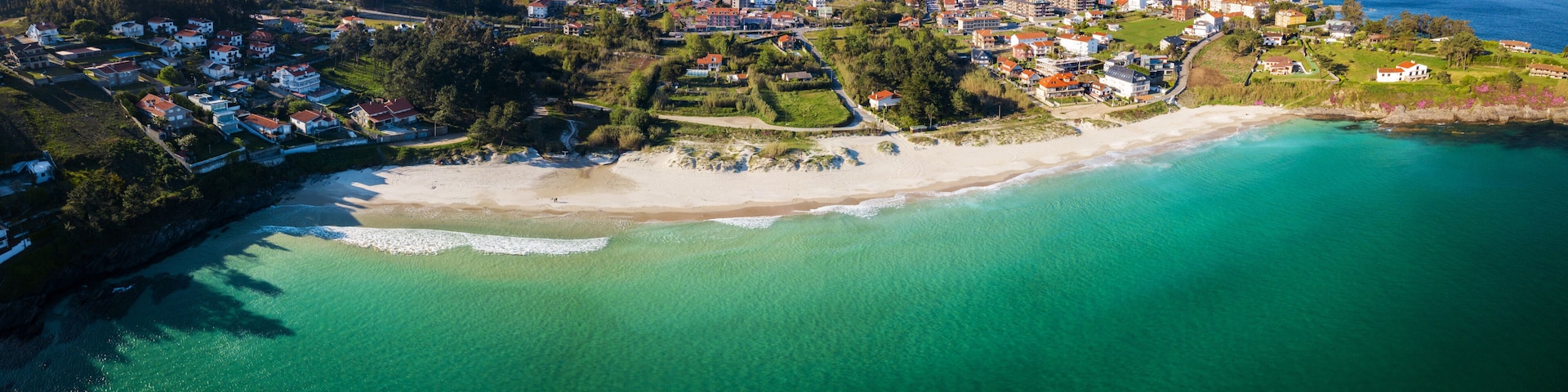 An aerial view of a coastal town with a sandy beach, clear turquoise waters, and a dense cluster of houses with red roofs.