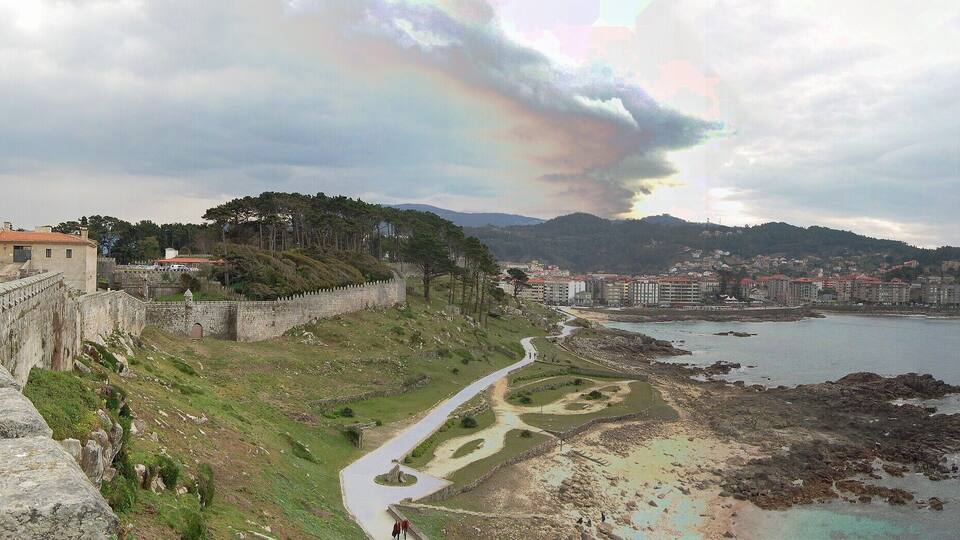 Una ciudad costera con un muro de piedra histórico, un sendero sinuoso y vistas al mar y montañas distantes.