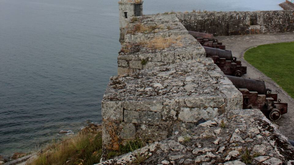 Una fortificación costera con cañones que dominan un mar tranquilo, una pequeña torre y colinas al fondo.