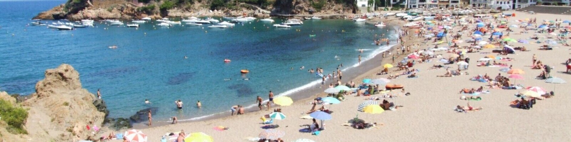 Praia lotada com pessoas se bronzeando e nadando, cercada por edifícios e barcos.