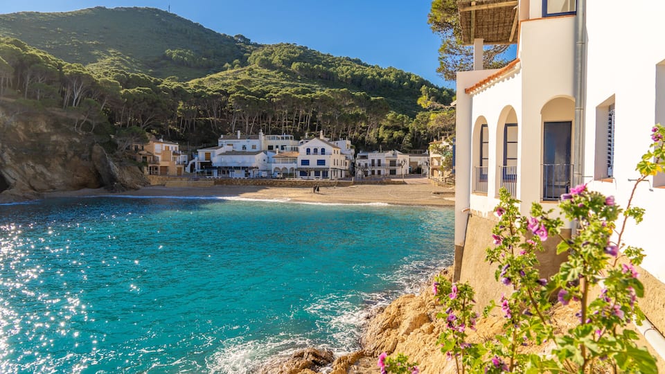 A coastal town with turquoise waters, white buildings, and a hillside in the background.