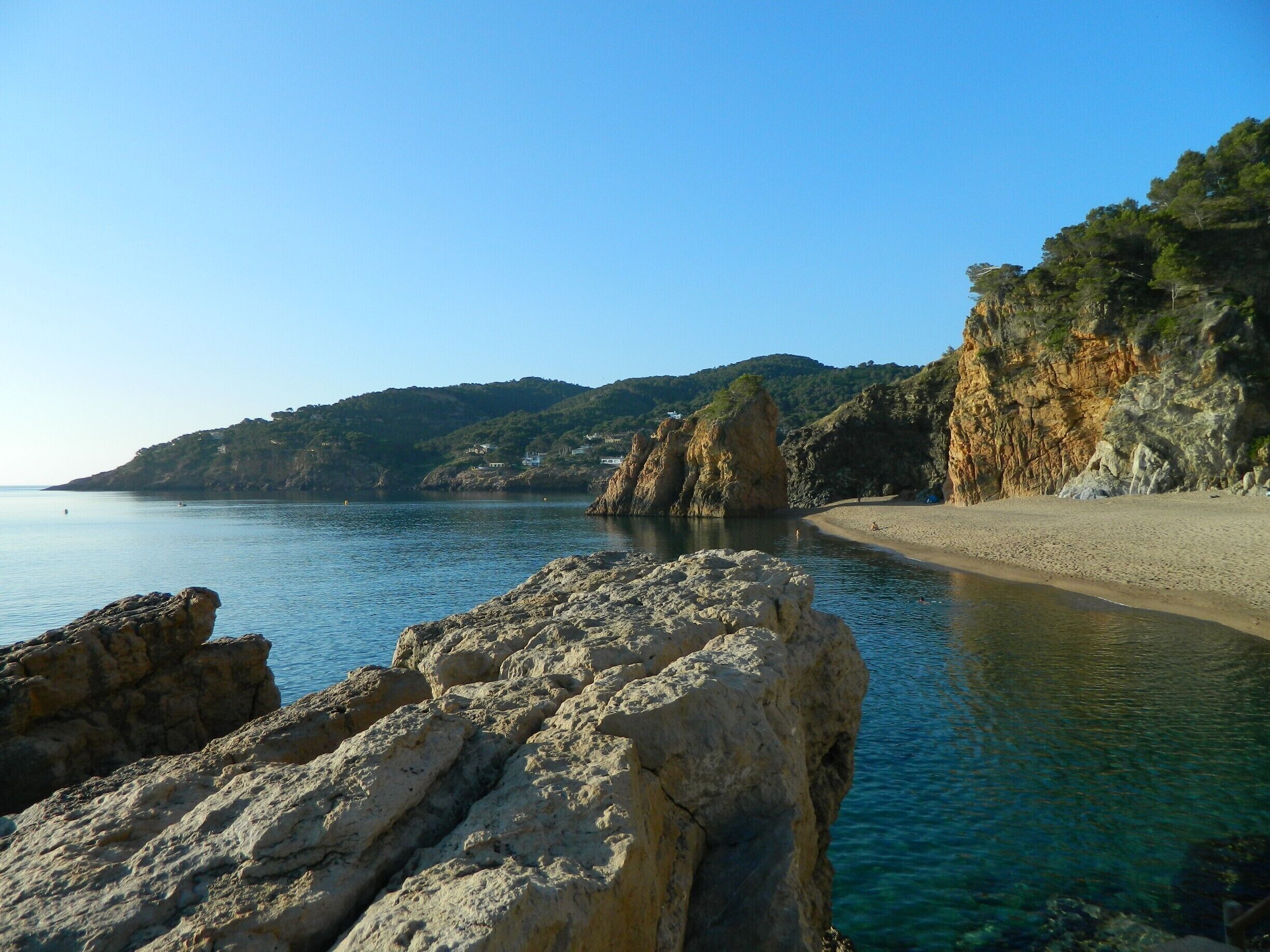A coastal landscape with clear blue water, rocky formations, and a sandy beach.