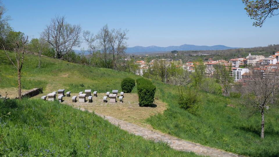 A grassy hillside with stone benches, a stone path, and a view of a town with buildings in the distance.