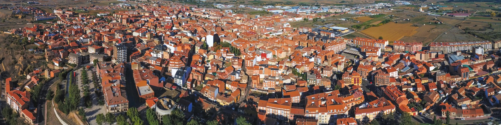 Una zona urbana densamente poblada con edificios de tejados rojos, rodeada de campos abiertos y colinas lejanas.
