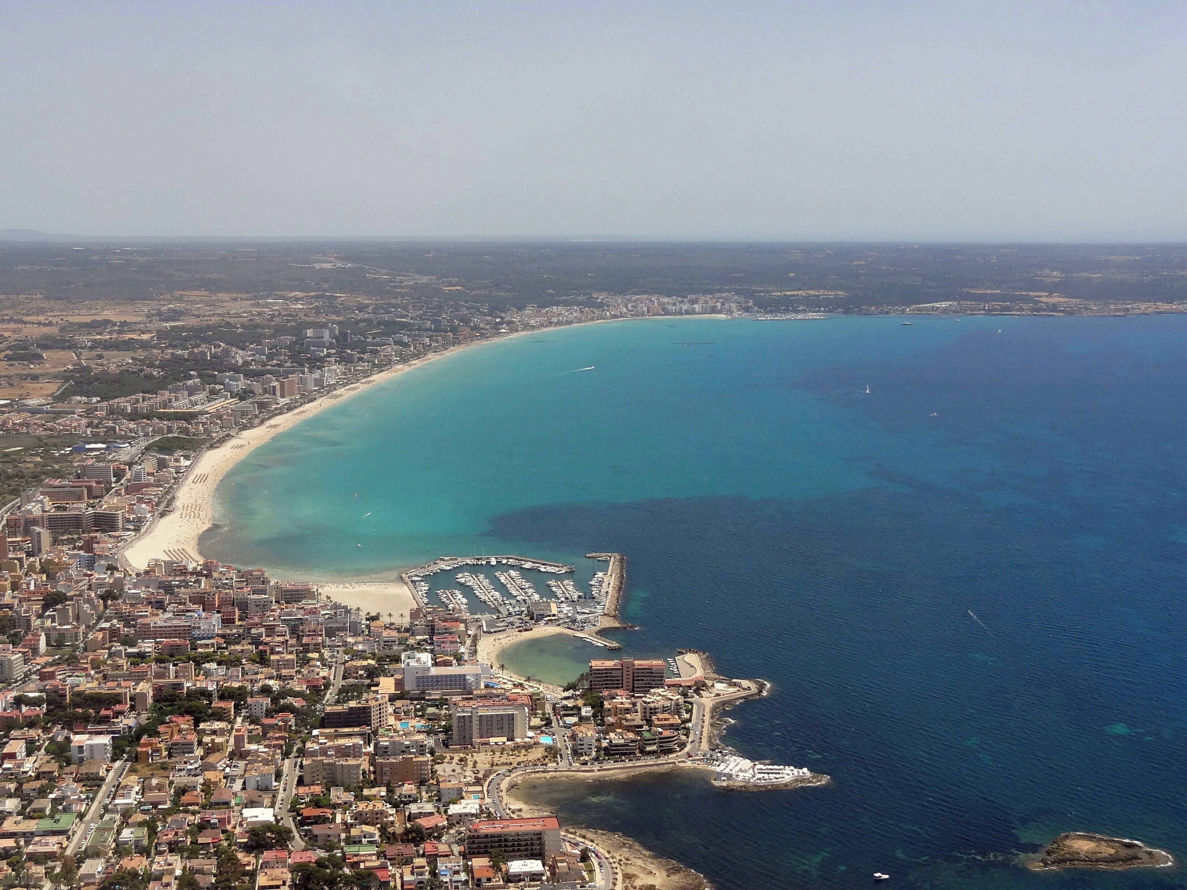 Vue aérienne d’une ville côtière avec une longue plage de sable, un port de plaisance et une zone urbaine densément construite.