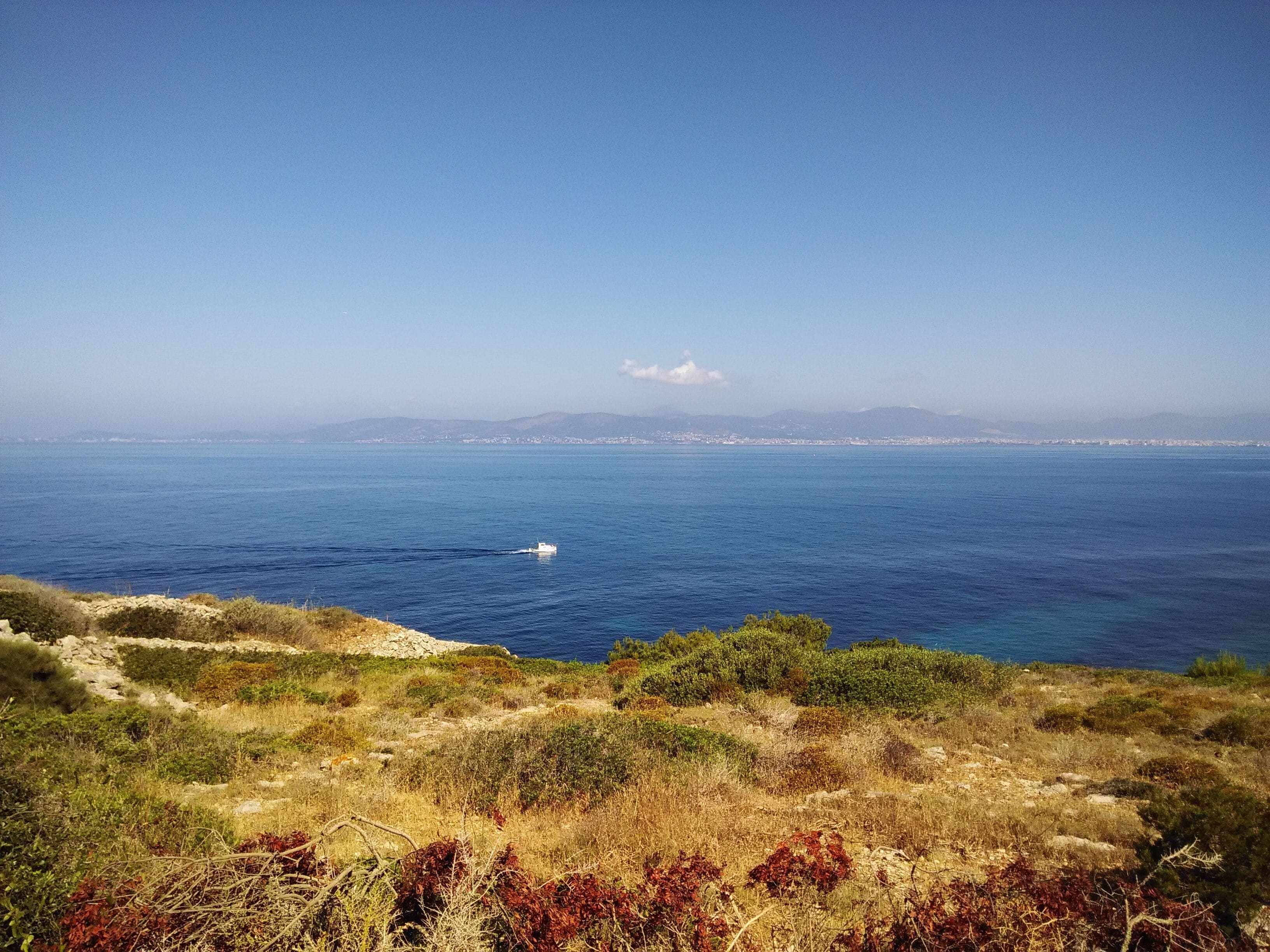 A coastal landscape with a boat on the sea, distant mountains, and a clear sky.