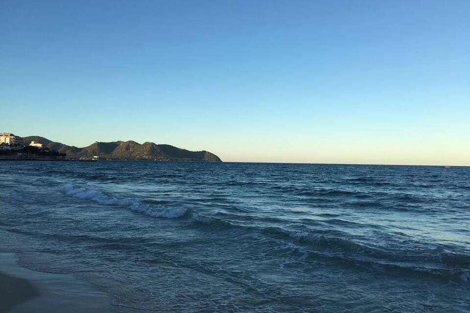 A coastal scene with waves, a distant mountain, and buildings along the shore.
