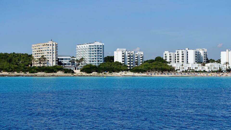 A coastal cityscape with a clear blue sea, white buildings, and a sandy beach.
