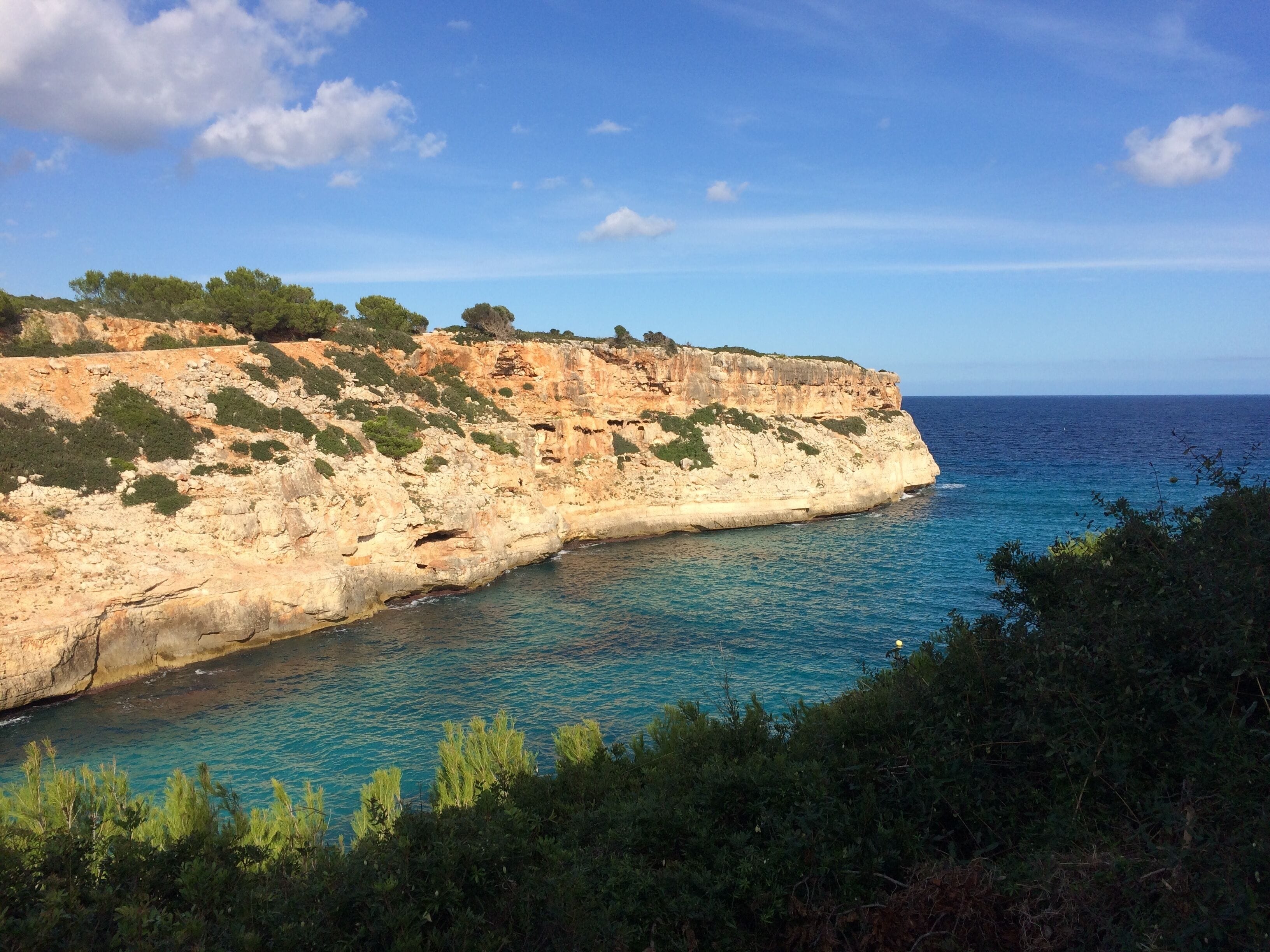 A coastal cliff with a clear blue sea, rocky terrain, and sparse vegetation.