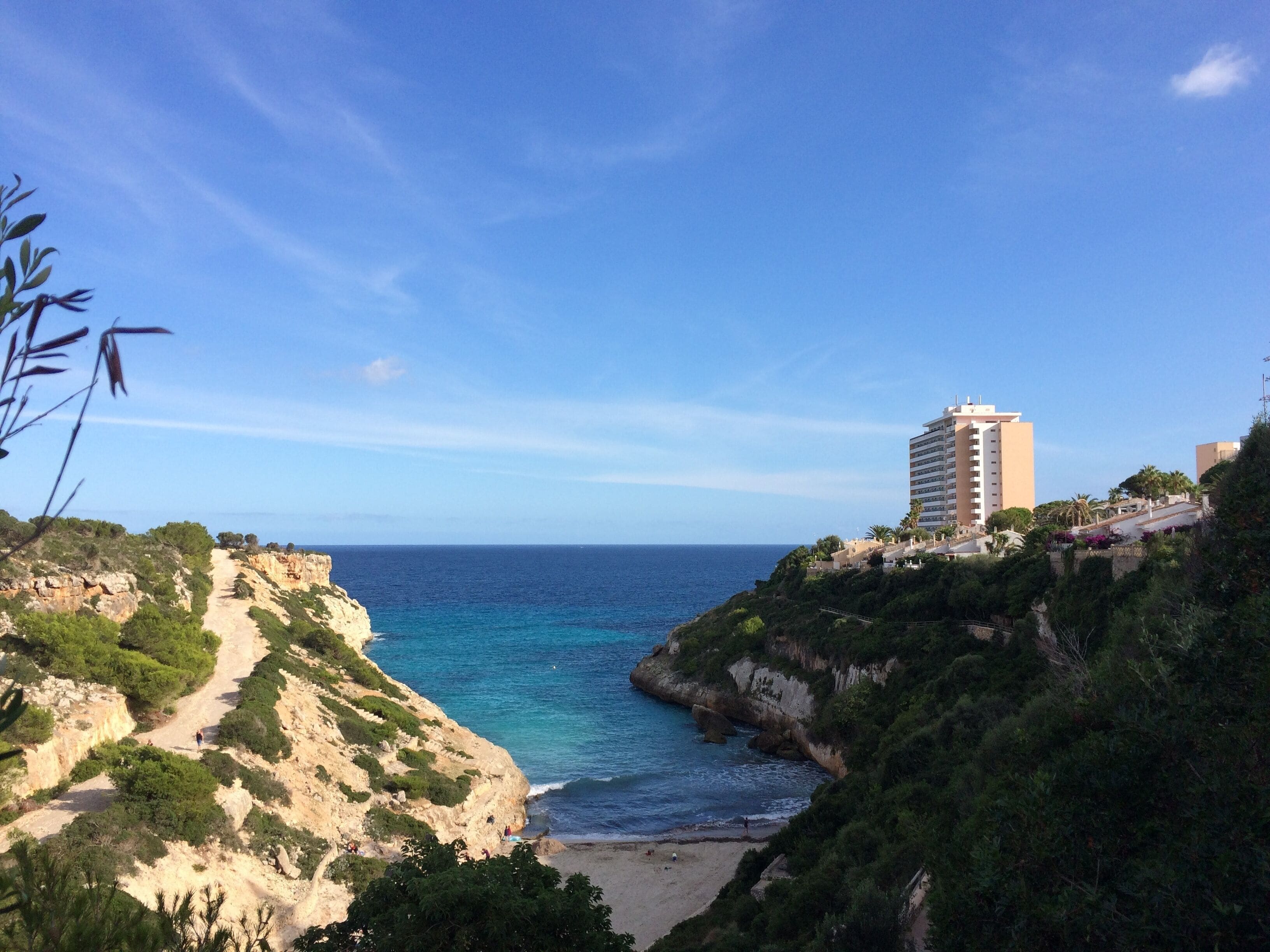 A coastal landscape with a clear blue sea, a sandy beach, rocky cliffs, and a tall building on a hill.