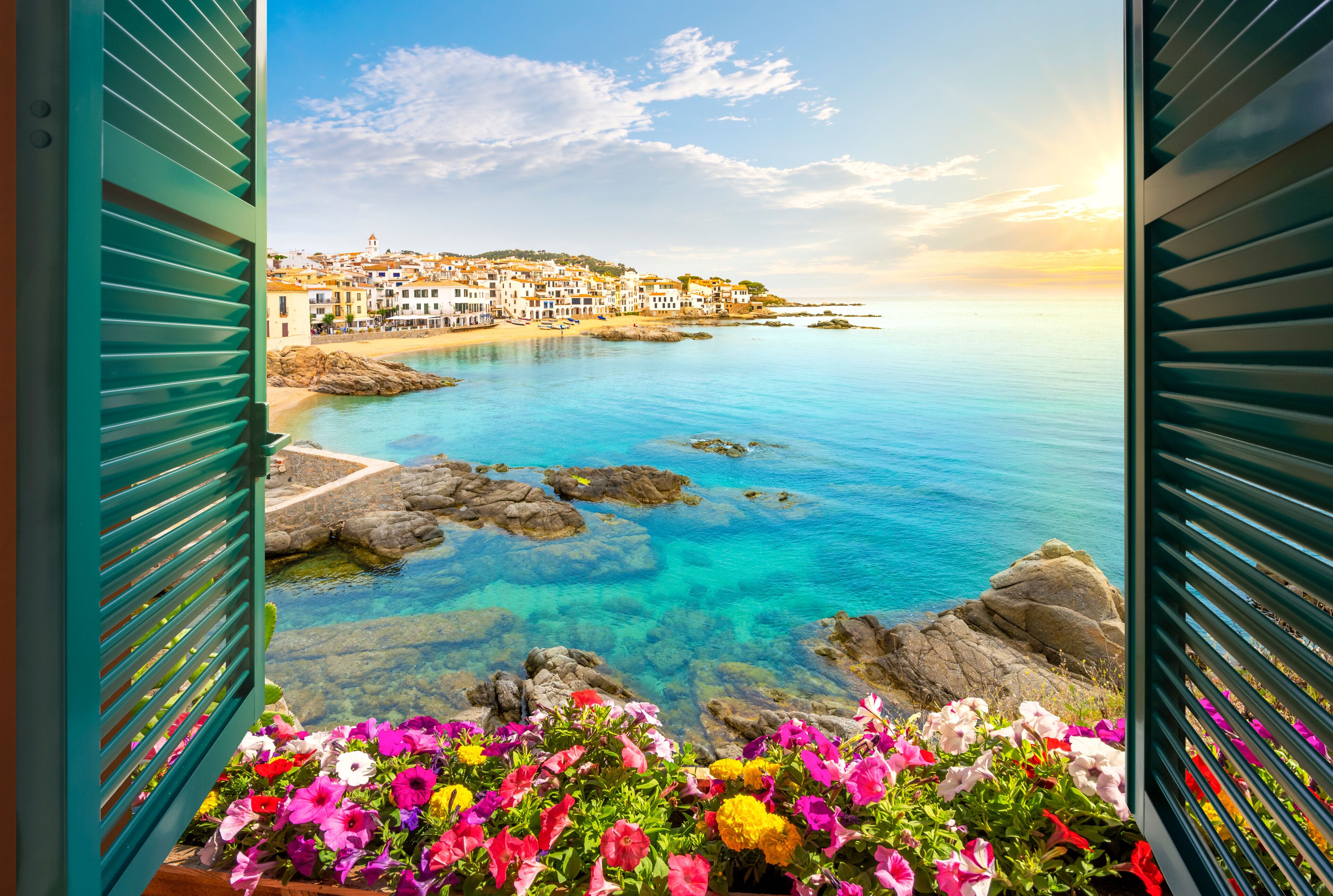 A coastal town with colorful flowers in the foreground, clear blue waters, and buildings on a hillside.