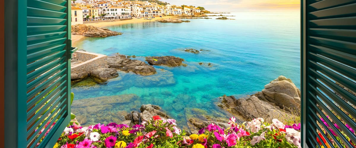 A coastal town with colorful flowers in the foreground, clear blue waters, and buildings on a hillside.