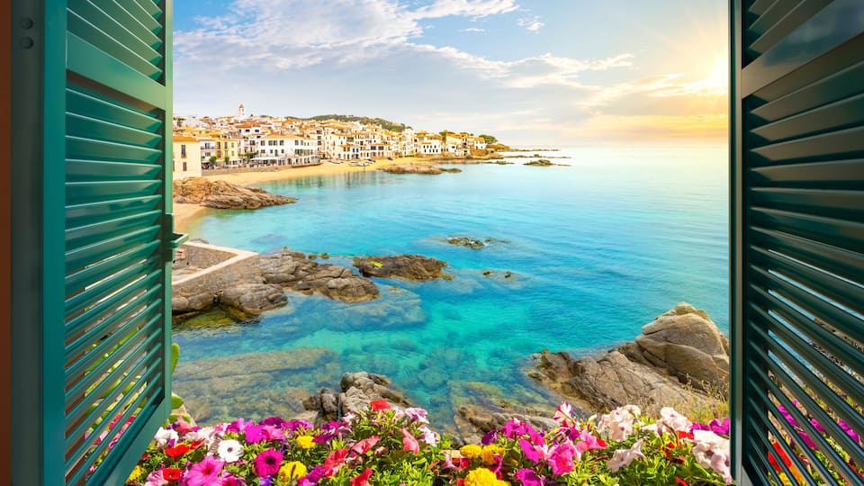 A coastal town with colorful flowers in the foreground, clear blue waters, and buildings on a hillside.