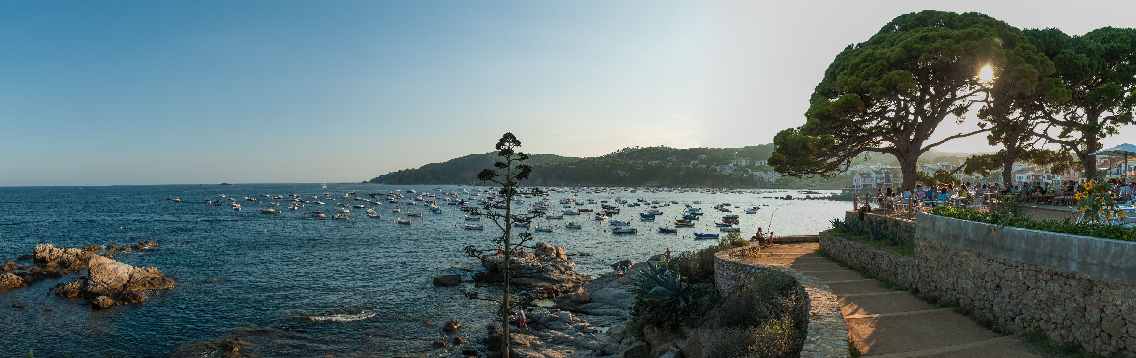 A coastal scene with a stone pathway, rocky shoreline, and numerous boats in the water.