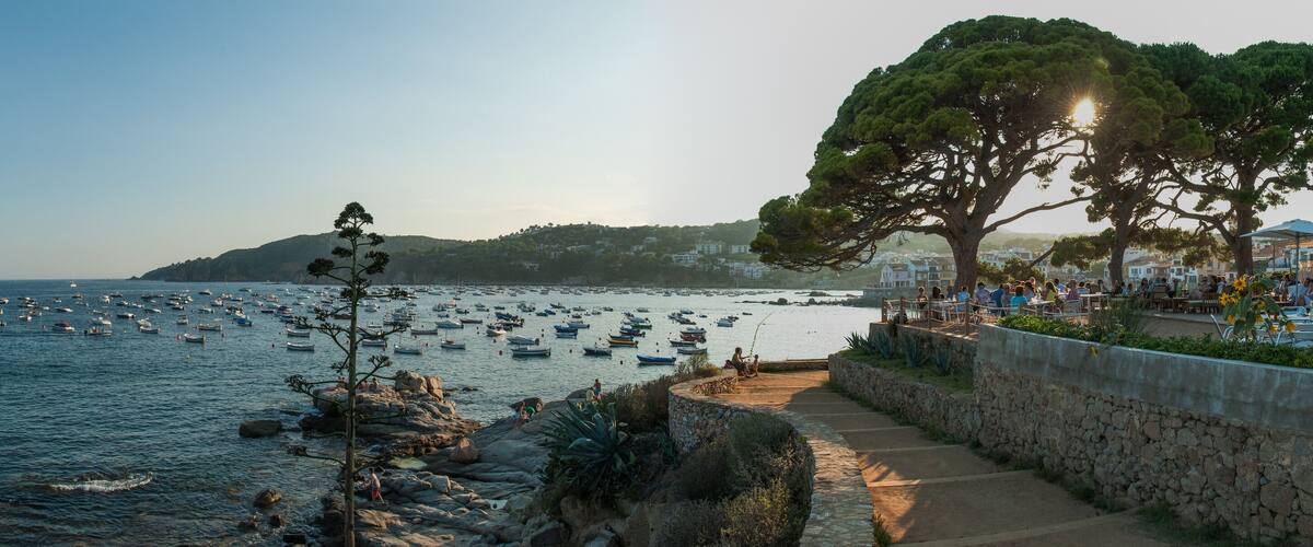 A coastal scene with a stone pathway, rocky shoreline, and numerous boats in the water.